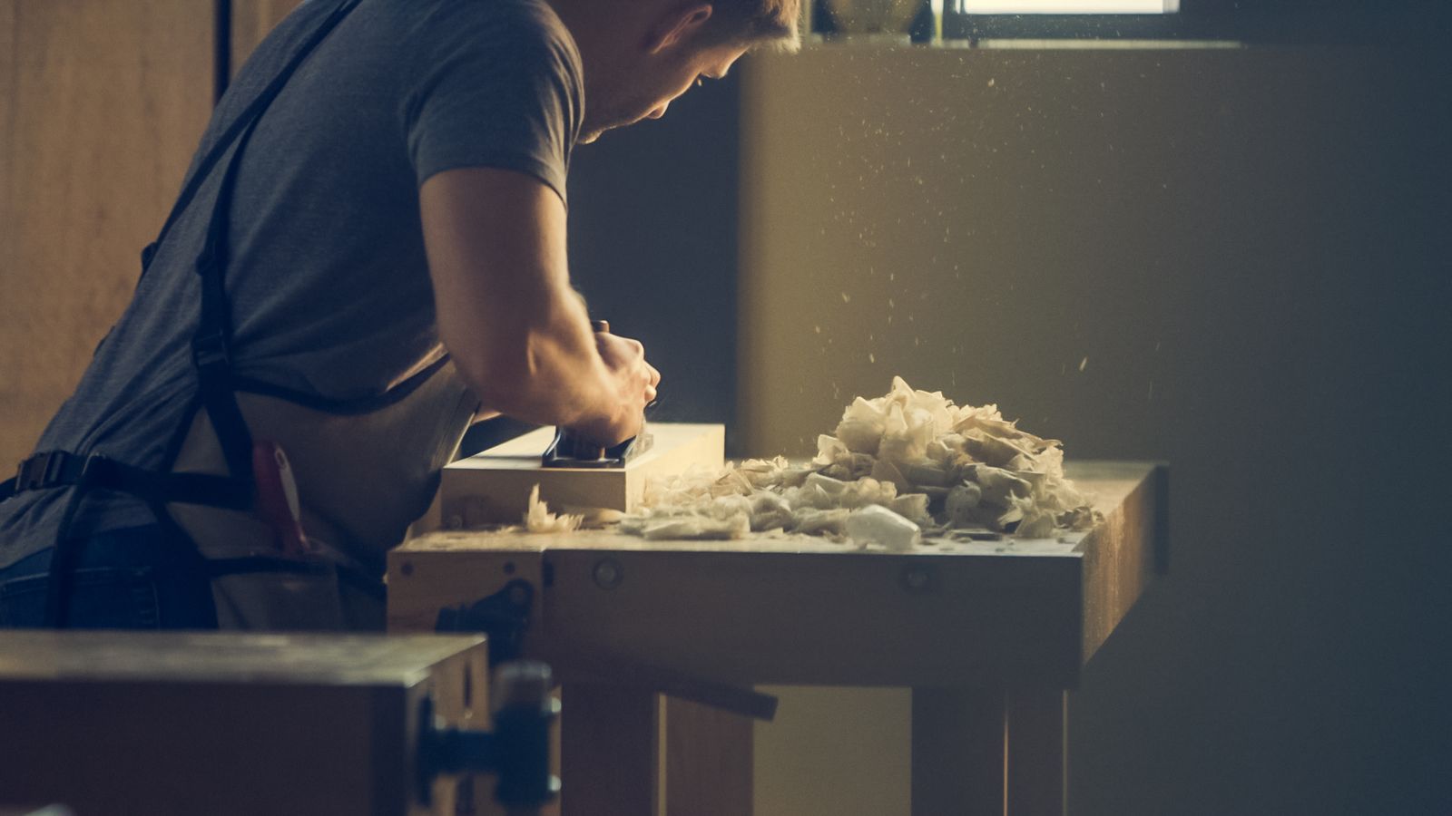 A carpenter hand-planing a board, fine shavings curling into warm light