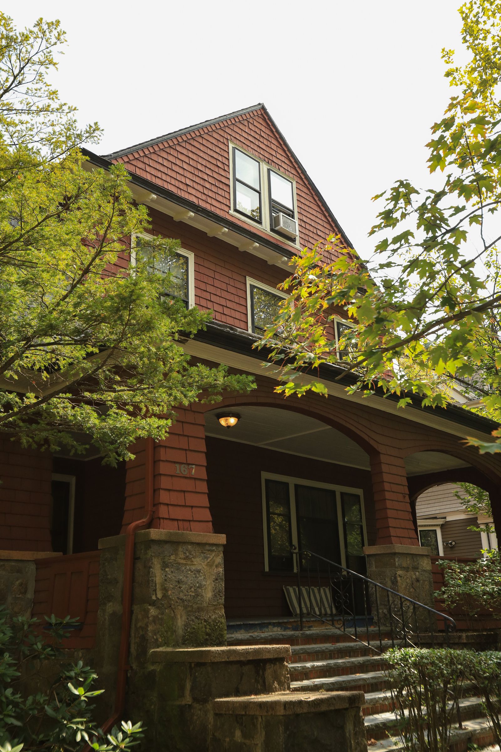 A stately maroon shingle-sided craftsman home with stone porch columns surrounded by lush greenery